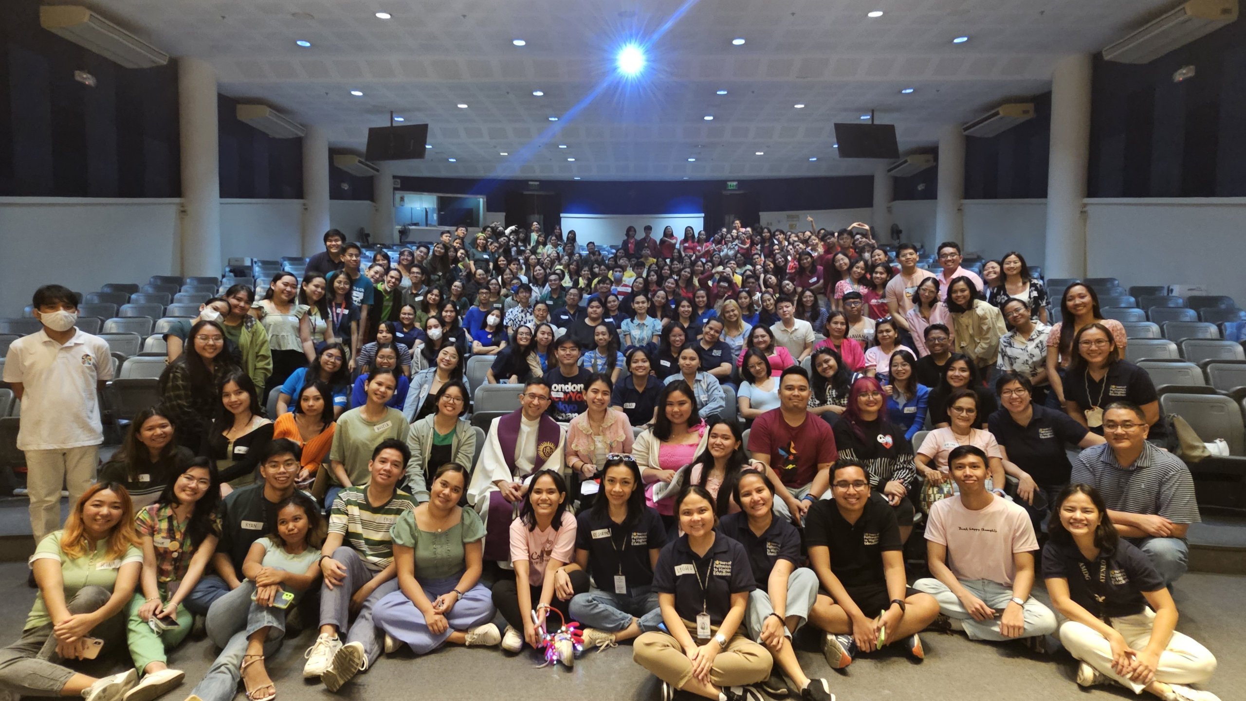 300 and more people, in a mix of students, professional alumni, and staff pose in an auditorium for a community photo. Everyone is smiling.