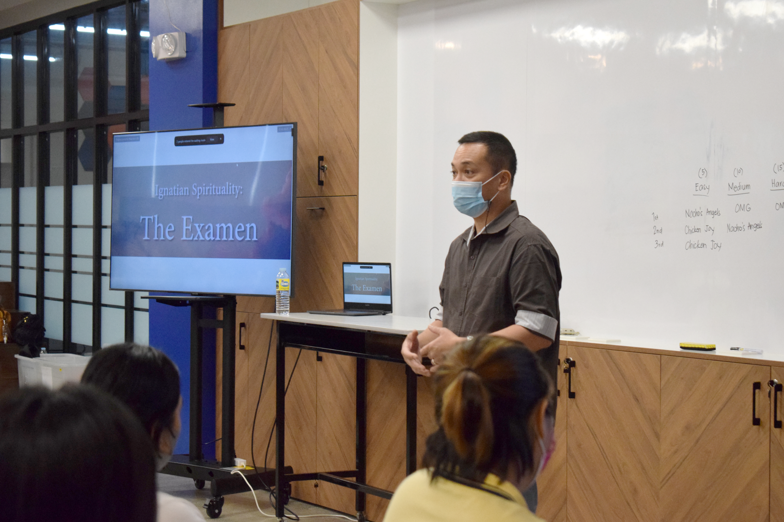 Volunteer Facilitator Cholo Santos from the Ateneo Campus Ministry Office leads the examen. In the background are a TV and monitor that display the word "Examen".