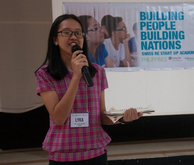 A young student presents an idea. She is holding a microphone and a block of pad paper.